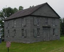 Vue générale de l'Église de Old Hay Bay, qui montre la masse architecturale à deux étages, en forme de boîte, rectangulaire, coiffée par un toit en pignon à pente moyenne;; Parks Canada Agency / Agence Parcs Canada, 2002.