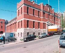 Vue en angle du Manège militaire, où l'on peut apercevoir les minces cordons de pierre taillée, le mur des fondations en pierre à taille éclatée et les échauguettes au-dessus de l’entrée, qui révèlent la vocation militaire du bâtiment, 1994.; Parks Canada Agency / Agence Parcs Canada, 1994.