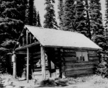 Vue en angle du Chalet des gardes du parc au lac Eva, qui montre ses murs en rondins posés à l’horizontale, assemblés à queue d’aronde et percés d’une porte en planches et de deux fenêtres à guillotine fixe à carreaux multiples, 1986.; Parks Canada Agency / Agence Parcs Canada, 1986.