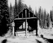 Vue de la façade du Chalet des gardes du parc au lac Eva, qui montre le pignon avant du chalet enchâssé dans un porche encadré par les pannes prolongées du toit et par les poteaux verticaux qui servent de support, 1986.; Parks Canada Agency / Agence Parcs Canada, 1986.