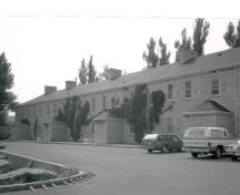 General view of the De Noyan Block, showing its domestic scale and simplicity of design consisting of a two-storey rectangular structure with a prominent gable-hipped roof with projecting stone chimneys, 1989.; (Environment Canada, Canadian Parks Service, Architectural History Branch/ Environnement Canada, Service canadien des parcs,  Direction de l'histoire de l'architecture, J. Adell, 1989.)