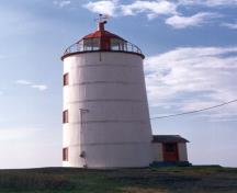 Vue latérale du Phare, montrant sa maçonnerie cylindrique, basse et légèrement effilée et son revêtement de planches blanches posées à la verticale et cerclées de cinq anneaux de métal, 1989.; Ministère des Affaires culturelles du Quebec / Quebec Ministry of Cultural Affairs, 1989.