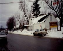 Vue de l'extérieur du magasin et entrepôt Saint-Laurent, qui montre le parement de planches à clin peint en blanc et le toit à deux versants dépourvu de lucarnes, 1993.; Parks Canada Agency / Agence Parcs Canada, 1993.