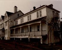 Corner view of Building 14, showing the simple, principal façade that features gables with false half timbering, a front porch, long verandahs and exterior fire escapes.; Agence Parcs Canada / Parks Canada Agency.