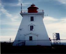 Vue latérale du phare, qui montre les détails d’inspiration classique de la corniche à consoles et l’alignement vertical des hautes fenêtres et de la porte en saillie surmontée de toits en appentis et à pignon, vers 1990.; Transport Canada / Transports Canada, circa / vers 1990.