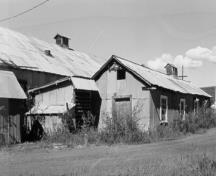 Corner view of the Tin Shop, showing the simple rectangular shape, gable roof, metal corrugated siding and roof covering, and the rooftop ventilator, 1988.; Agence Parcs Canada / Parks Canada Agency, 1988.