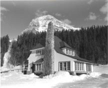 Façade latérale de la résidence du directeur, qui montre le toit en croupe très pentu et complétée par une cheminée en pierres des champs placées de façon irrégulière, vers 1965.; Agence Parcs Canada / Parks Canada Agency, ca./vers 1965.
