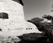 Side view of Cathcart Tower, showing the massively constructed, smooth, steep exterior walls built of tightly fitting limestone blocks and two of the four evenly spaced caponiers around the base, 1992.; Parks Canada Agency / Agence Parcs Canada, 1992.