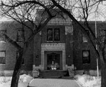 Front elevation of the Yorkton Armoury, showing the stone detailing of the main entrance, including the articulated Art Deco geometry on the surrounds, and the sculpted panels depicting heavy artillery, 1989.; Department of National Defence / Ministère de la Défense nationale, 1989.