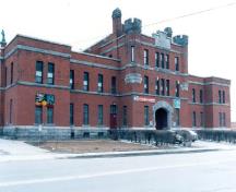 Vue de la façade avant du Manège militaire montrant les éléments empruntés aux ouvrages militaires de style médiéval, y compris les tourelles crénelées, les cheminées et la grande porte, 1989.; Department of National Defence / Ministère de la Défense nationale, 1989