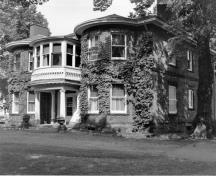 Vue de coin de la villa Fairholm démontrant les murs de briques rouges tachetées; et la pierre de taille utilisée pour les linteaux au-dessus des fenêtres, pour le sous-sol et comme ceinture entre les deux étages.; Parks Canada Agency / Agence Parcs Canada, n.d.
