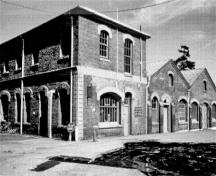 General view of building D51's main (western) and side (northern) elevations showing the red brick walls, stone dressings, corbelled friezes, vaults and arches, 1989.; Parks Canada Agency / Agence Parcs Canada, I. Doull, 1989.