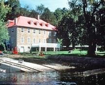 Corner view of the river side of the Carillon Barracks showing the location of the building on the banks of the Ottawa River, next to the Carillon Canal, 1989.; Parks Canada Agency/ Agence Parcs Canada, 1989.