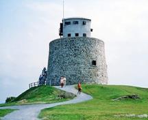 Vue générale ce la tour martello de Carleton montrant les murs construits en moellons bruts, qui s’inclinent légèrement vers l’intérieur à mesure qu’ils s’élèvent jusqu’au mur du parapet surmonté par un poste d’observation en béton de deux étages, 1983.; Parks Canada Agency / Agence Parcs Canada, J. Steeves, 1983.