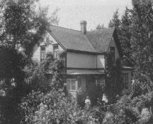 Exterior view of Building D1, showing the asymmetrical massing of the L-shaped, two-storey structure with its steep gable roof and chimney, 1971.; CIHB/IBHC, Parks Canada/Parcs Canada, 1971