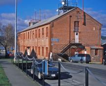 General view of Building D77 Former Ordnance Stores, showing the symmetrical massing of this two-storey, rectangular, gable-roofed building.; Parks Canada Agency / Agence Parcs Canada, Ian Doull.