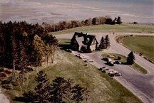 Vue aérienne du centre d’information aux visiteurs Est, qui montre la conception pittoresque du bâtiment et son emplacement dans un endroit dégagé, qui fait ressortir la nature rustique du cadre, vers 1980.; Parks Canada Agency / Agence Parcs Canada, c./vers 1980.